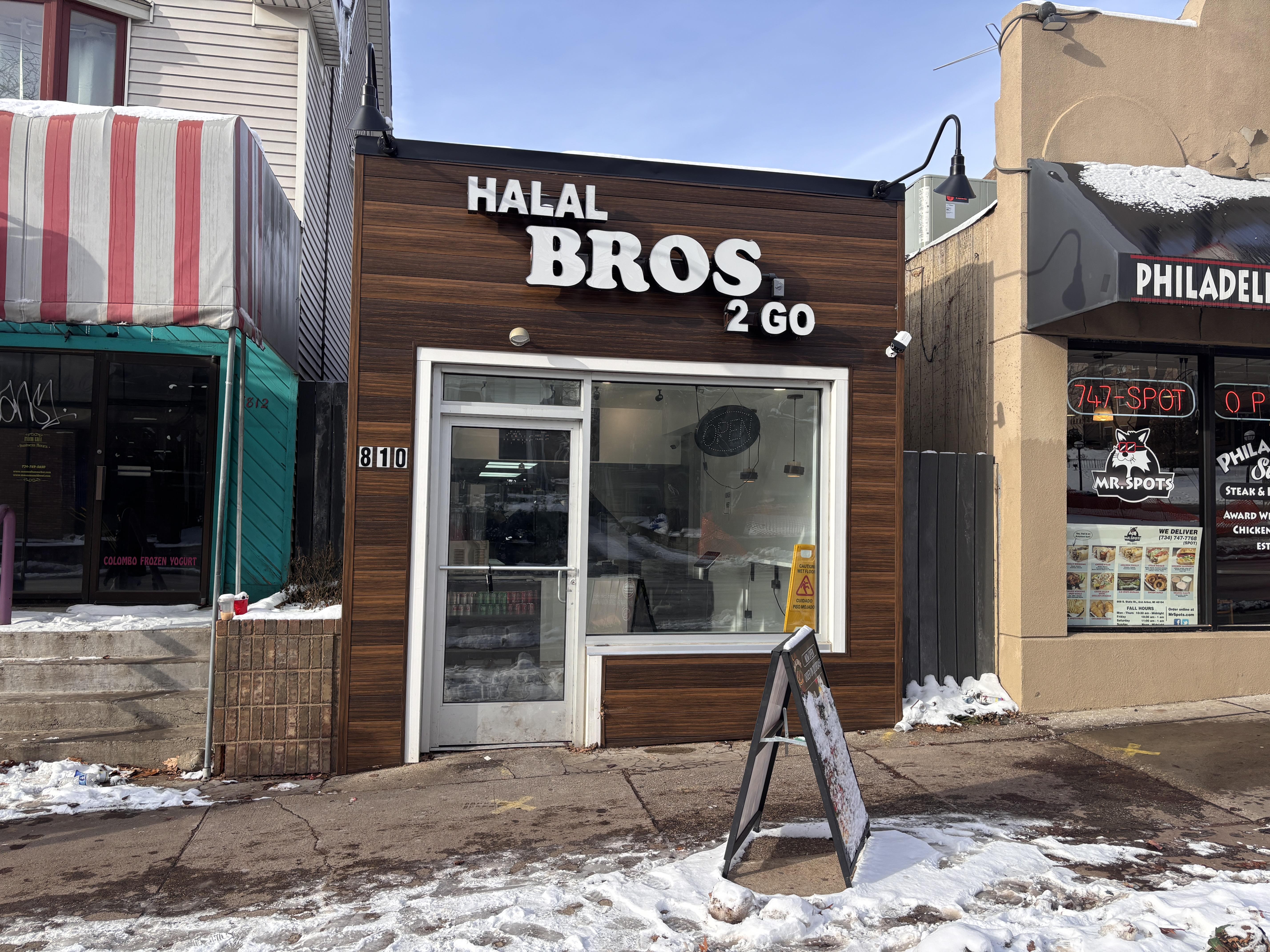 Exterior view of a restaurant named 'Halal Bros 2 Go' featuring a modern facade, with a nearby colorful storefront and snow on the ground.